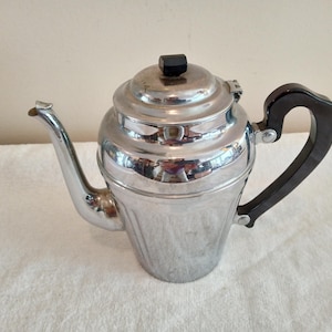 May include: A vintage silver-colored teapot with a black handle and lid knob. The teapot has a curved spout and a rounded body. The handle is a dark, curved shape. The teapot is sitting on a white surface.