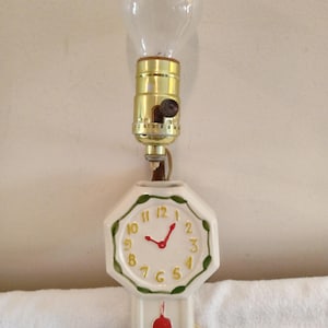 May include: A unique lamp featuring a clear light bulb above a white ceramic clock. The clock has a red pendulum and hands, with gold numerals and a green border. The lamp is plugged into a wall outlet.