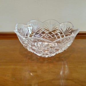 May include: Clear crystal glass bowl with a scalloped rim. The bowl features a cut glass design with diamond and fan patterns. The bowl is sitting on a wooden surface.