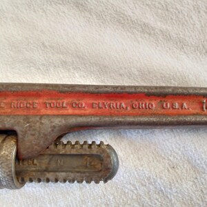 Op de afbeelding: Een vintage pijpsleutel van 35,56 cm van The Ridge Tool Co. in Elyria, Ohio, VS. De sleutel is voornamelijk roestig oranje met zichtbare tekst en een getande bek voor het vastgrijpen van pijpen. De tool vertoont slijtage.