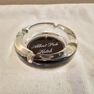 May include: Clear glass ashtray with a black interior base. The ashtray has a rounded shape with four resting points for cigarettes. The text "Albert Pick Hotels" is printed in white script on the base.