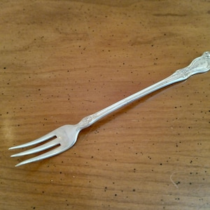 May include: A silver fork with three tines, resting on a wooden surface. The handle has an ornate design with a decorative flourish at the end. The fork is likely vintage or antique.