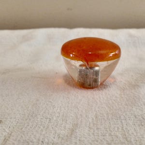 May include: A clear, teardrop-shaped bottle stopper with an amber-colored top. The stopper has a silver-colored, ridged base. The stopper is sitting on a white, textured surface.