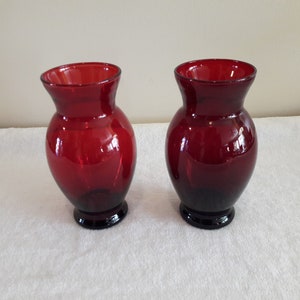 May include: Two red glass vases with a rounded body and a flared top. The vases are on a white surface.