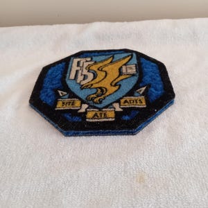 May include: An octagonal embroidered patch featuring a blue shield with a gold eagle emblem. The patch includes white lettering "FS" and three yellow banners with the text "FITE", "ATE", and "ADTS" set against a dark blue and black background.