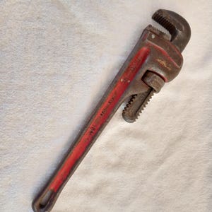 Op de afbeelding: Een vintage pijptang met een rode handgreep en een donker metalen kop. De tang is ongeveer 35,5 cm lang en vertoont tekenen van slijtage. De verstelbare bekken staan open, klaar voor gebruik.