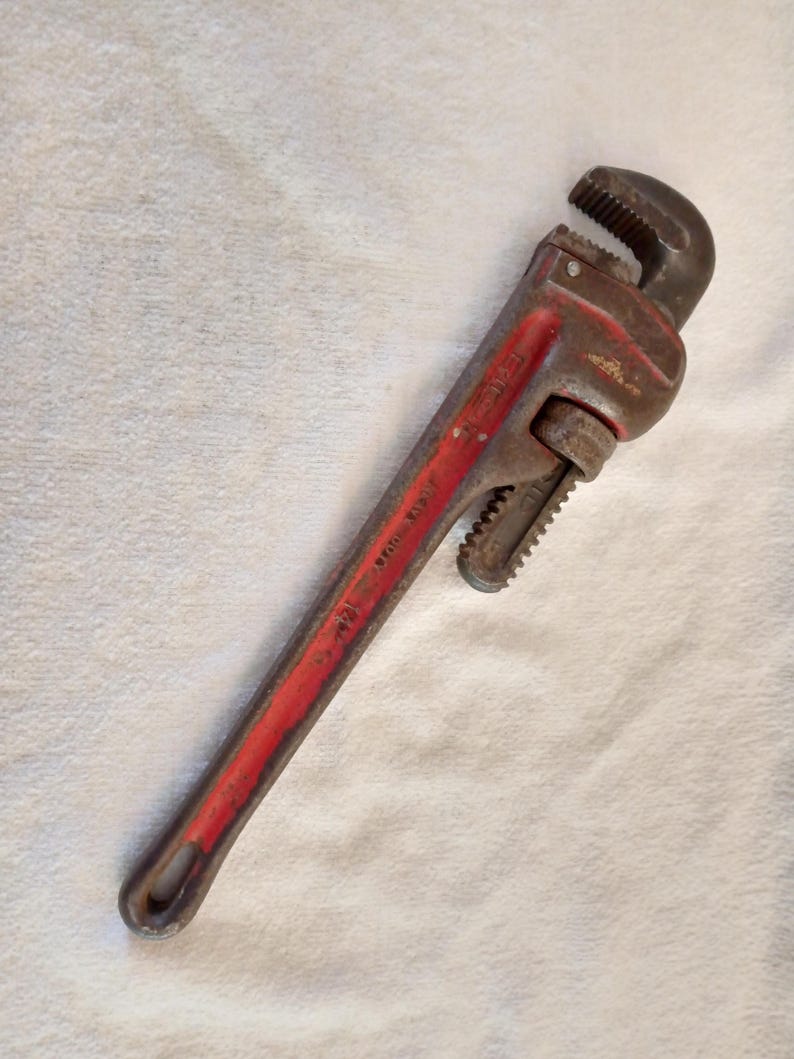 Op de afbeelding: Een vintage pijptang met een rode handgreep en een donker metalen kop. De tang is ongeveer 35,5 cm lang en vertoont tekenen van slijtage. De verstelbare bekken staan open, klaar voor gebruik.