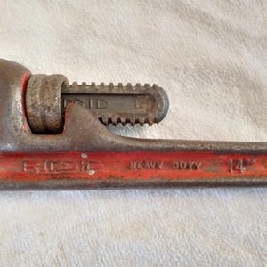 Op de afbeelding: Een vintage, heavy-duty RIDGID pijpsleutel, 35,56 cm lang, met een rode handgreep en een roestige, versleten uitstraling. De sleutel is ontworpen voor het vastgrijpen en draaien van pijpen en andere cilindrische objecten.