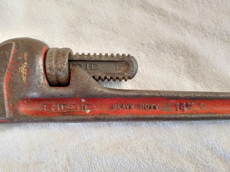 Op de afbeelding: Een vintage, heavy-duty RIDGID pijpsleutel, 35,56 cm lang, met een rode handgreep en een roestige, versleten uitstraling. De sleutel is ontworpen voor het vastgrijpen en draaien van pijpen en andere cilindrische objecten.