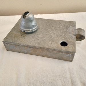 May include: A vintage metal object with a bell-shaped top and a rectangular base. The object has a small ring attached to the bell. The base has a circular hole and a curved handle-like extension. The metal has a weathered, aged appearance.
