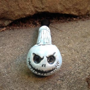 May include: A ceramic bottle stopper featuring a Jack Skellington design. The stopper has a white base with a black, stitched mouth and eye details. The top portion is light blue with vertical lines and a black knob.