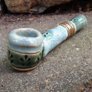 May include: A ceramic smoking pipe featuring a light blue crackle glaze finish on the bowl and stem. The bowl base is decorated with a dark green band containing a repeating leaf pattern, while the stem includes a textured copper-colored ring.