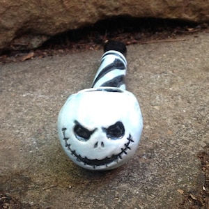 May include: A ceramic pipe featuring a Jack Skellington face design. The bowl is white with black eyes and a stitched mouth. The stem is black and white striped. The pipe is resting on a gray stone surface.