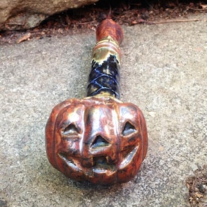 May include: A ceramic Halloween-themed pipe. The bowl is shaped like a carved pumpkin with a smiling face. The stem is a mix of colors, including brown, blue, and green. The pipe is designed for smoking.