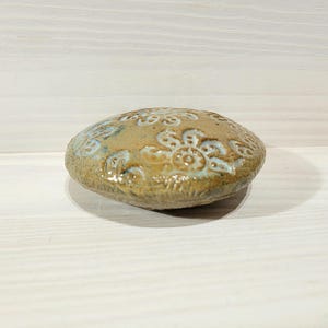 May include: A round, flattened ceramic object with a glossy, tan-coloured glaze. The surface features a repeating, embossed floral pattern with light blue accents within the recessed areas of the design, creating a textured, decorative appearance.