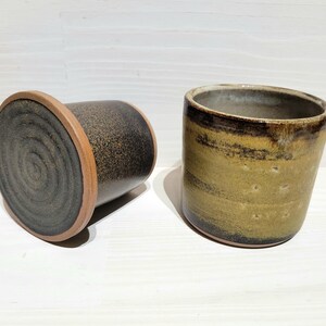 May include: Two cylindrical ceramic cups with a speckled finish. One cup is positioned on its side, revealing a dark grey base with a circular spiral pattern and a terracotta rim. The other cup stands upright, displaying a mottled olive-green glaze.