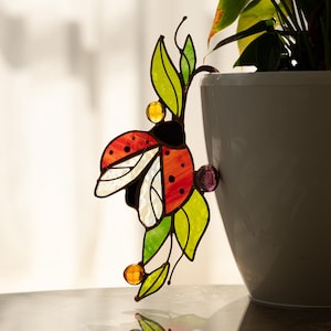 Op de afbeelding: Een gebrandschilderde lieveheersbeestjesdecoratie met rode, zwarte en witte vleugels en een groen bladerontwerp. Het lieveheersbeestje is geaccentueerd met gele en paarse glazen kralen. De decoratie is geplaatst in de buurt van een potplant.