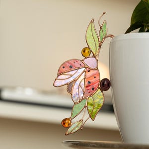 May include: A colorful stained glass plant stake in the shape of a butterfly. The butterfly has pink, orange, and white wings with black dots. The leaves are green and the accents are amber and purple. The stake is next to a white pot.