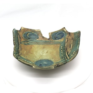 May include: A ceramic bowl with an abstract design in shades of brown, green, and blue. The bowl has a slightly irregular shape and a chipped edge.