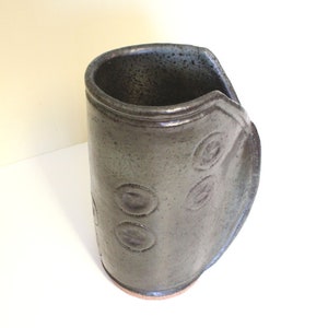 May include: A dark green ceramic vase with a unique, slightly curved shape. The vase has a textured surface and a series of circular indentations.