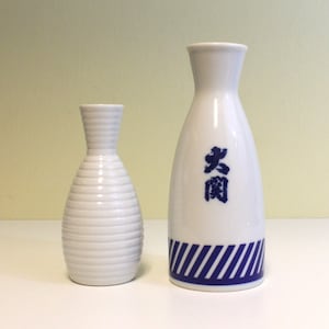 May include: Two white ceramic sake bottles. The smaller bottle has a ribbed design. The larger bottle has a blue and white striped design and the word "老酒" in blue.