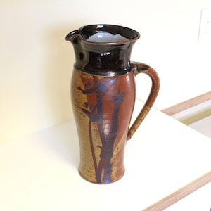 May include: A brown and black ceramic pitcher with a stylized design. The pitcher has a wide mouth and a handle.
