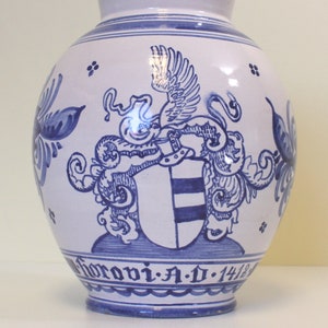 May include: A white ceramic vase with blue and white painted designs. The vase features a coat of arms with a winged creature and the inscription "Horovia A.D. 1412".