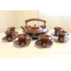 May include: A brown and black ceramic teapot with a wooden handle and six matching cups and saucers. The cups and saucers have a triangular shape and a speckled design.