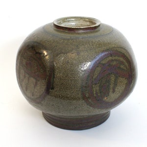 May include: A round, brown ceramic vase with a textured surface and a dark brown glaze. The vase has a wide base and a narrow neck. The vase is decorated with a pattern of circles and lines in a darker brown glaze.