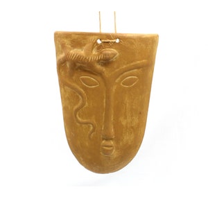 May include: A terracotta wall hanging in the shape of a face with a lizard on top. The face has a long nose and closed eyes. The hanging has a rustic, earthy tone.
