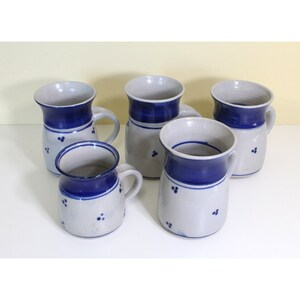 May include: Five ceramic mugs with blue and white designs. The mugs have a rounded body and a handle. The mugs are decorated with a blue band around the top and a white background with blue dots.