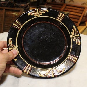 May include: A black ceramic plate with a brown rim and a decorative design of stylized yellow and brown floral patterns around the edge.