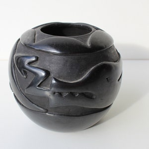 May include: Black pottery vase with a geometric design. The vase is round and has a wide opening. The design is carved into the surface of the vase and includes a lightning bolt and a bird.