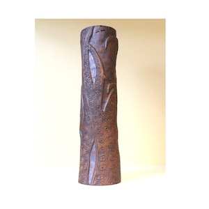 May include: A tall, cylindrical wooden vase with a carved design of swirling leaves and circles. The vase is a dark brown color and has a rough, textured surface.
