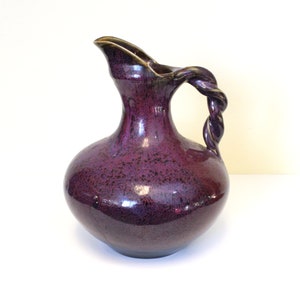 May include: A purple ceramic pitcher with a twisted handle. The pitcher has a speckled glaze and a glossy finish.