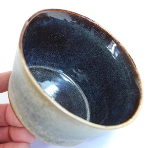 May include: A handmade ceramic bowl with a dark blue interior and a light gray exterior. The rim is a dark brown color. The bowl has a smooth, glossy finish and is likely used for serving food or as a decorative piece.
