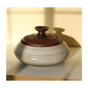 May include: A white ceramic lidded bowl with a brown lid. The lid has a raised center and a decorative pattern of lines. The bowl has a rounded shape and a smooth, white glaze.