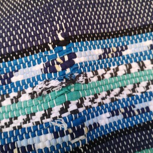 May include: A close-up of a woven rug with a blue, green, white, and black pattern. The rug is made of recycled materials and has a frayed edge.