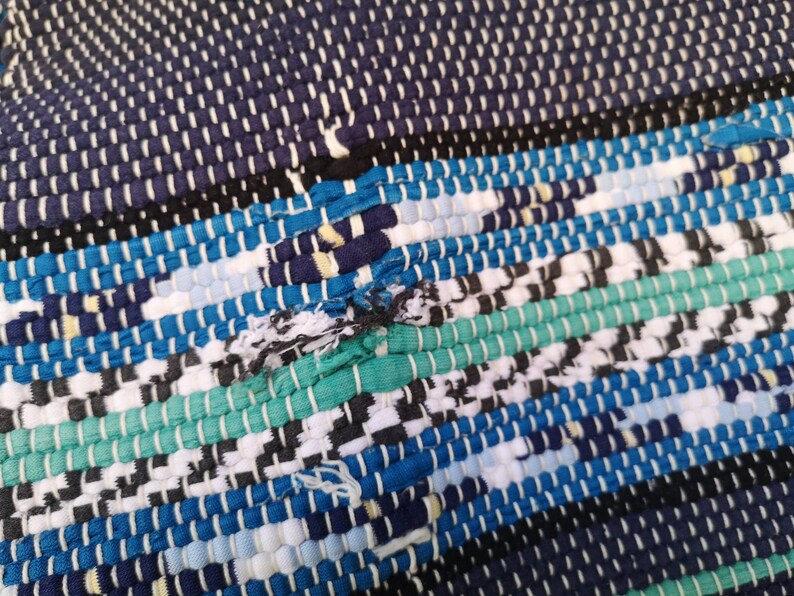 May include: A close-up of a woven rug with a blue, green, white, and black pattern. The rug is made of recycled materials and has a frayed edge.