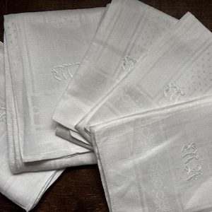 Puede incluir: Un montón de cinco servilletas de lino blanco con iniciales bordadas. Las servilletas están dobladas por la mitad y tienen una textura sutil.