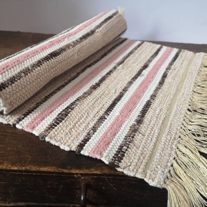 May include: A woven rug with pink and brown stripes and a fringe on the ends. The rug is rolled up and lying on a wooden surface.