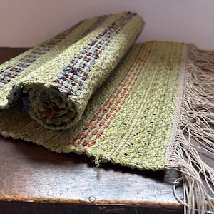 Könnte beinhalten: Handgewebter grüner und mehrfarbiger Flickenteppich mit einem gerollten Abschnitt. Der Teppich hat Streifen in Rot, Blau und Braun. Der Teppich hat an einem Ende einen Fransen. Der Teppich besteht aus gewebtem Stoff.
