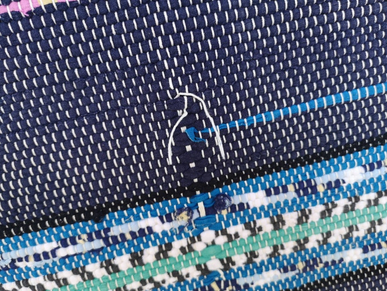 May include: Close-up of a blue and white woven rug with a white thread detail. The rug has a geometric pattern with blue, white, and green stripes.