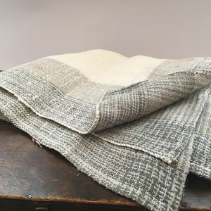 Könnte beinhalten: Gefaltetes gewebtes Textil in Creme-, Grau- und Beigetönen. Der Stoff hat ein strukturiertes Aussehen mit einem sichtbaren Webmuster. Die Kanten sind mit einem kontrastierenden Rand versehen, der über einer dunklen Holzoberfläche drapiert ist.