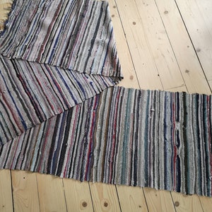 Pode incluir: Um tapete longo e estreito, tecido à mão, com um padrão listrado em tons de cinza, branco, marrom, azul e vermelho. O tapete é feito de materiais reciclados e tem um visual rústico e vintage.