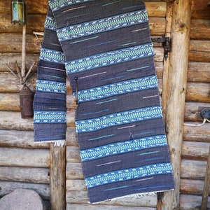 May include: A dark blue and green woven rug with a geometric pattern hangs on a wooden wall. The rug has a fringe on the bottom.
