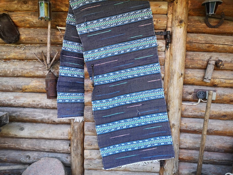 May include: A dark blue and green woven rug with a geometric pattern hangs on a wooden wall. The rug has a fringe on the bottom.