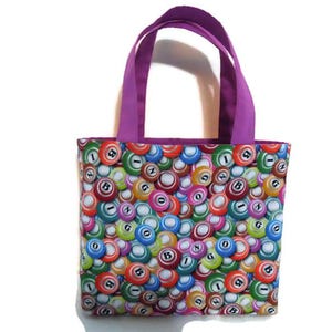 May include: A rectangular tote bag with a colorful pattern of bingo balls. The bag has a purple handle and trim. The bingo balls are in various colors, including red, green, blue, and yellow.