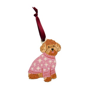 Op de afbeelding: Houten hondenornament met een roze trui met witte sneeuwvlokpatronen. De hond heeft een lichtbruine vacht en draagt een roze trui met witte sneeuwvlokpatronen. Een bordeauxrood lint is bevestigd om op te hangen.