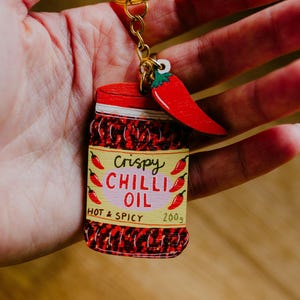 May include: A novelty wooden keyring depicting a jar of crispy chilli oil. The jar is red with the text "Crispy Chilli Oil" and "Hot & Spicy". A red chilli pepper charm is attached to the keyring.